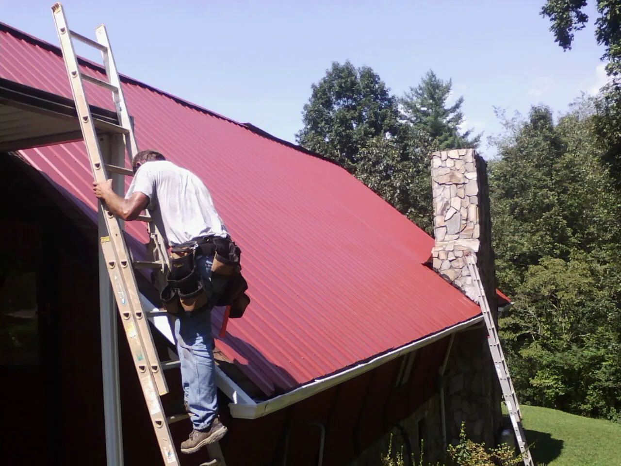 Roofer on a ladder beside a red metal roof and stone chimney — Soffit & Fascia Repair by Roofing Brothers in Point Pleasant